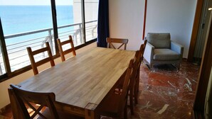 Een eethoek met een houten tafel en stoelen, uitzicht op de oceaan door grote ramen, en een marmeren vloer.