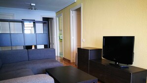 Een woonkamer met een grijze bank, een zwarte salontafel, een televisie op een houten standaard, en een keuken op de achtergrond.