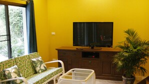 Ein Wohnzimmer mit gelben Wänden, einem Flachbildfernseher, Korbmöbeln und einem Topfpflanzen.