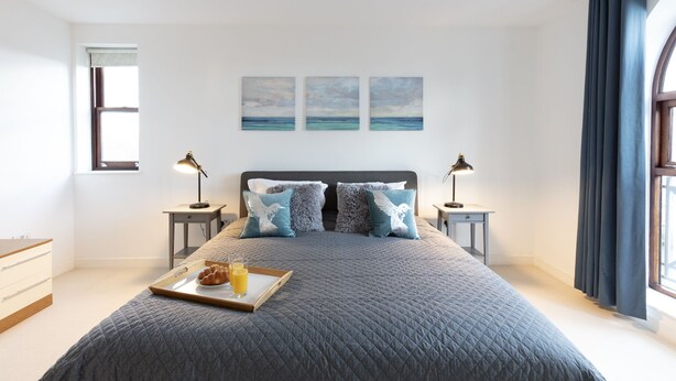 Une chambre moderne avec un grand lit, deux tables de chevet avec des lampes, un plateau avec des croissants et du jus de fruits, et une vue sur la mer par la fenêtre.