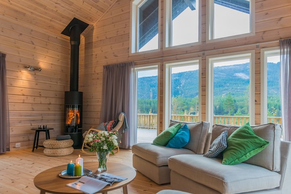 A cozy cabin interior with a wood-burning stove, a sofa with colorful cushions, a dining table with candles, and large windows offering a mountain view.