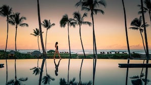 Eine Frau geht an einem Pool entlang, Palmen und Sonnenuntergang im Blick.