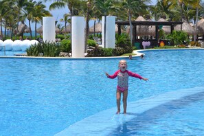 A pool area with a child standing on the edge, palm trees, and thatched-roof structures in the background. - Beautiful property    huge pool  best golf course in the area! (playa del carmen)