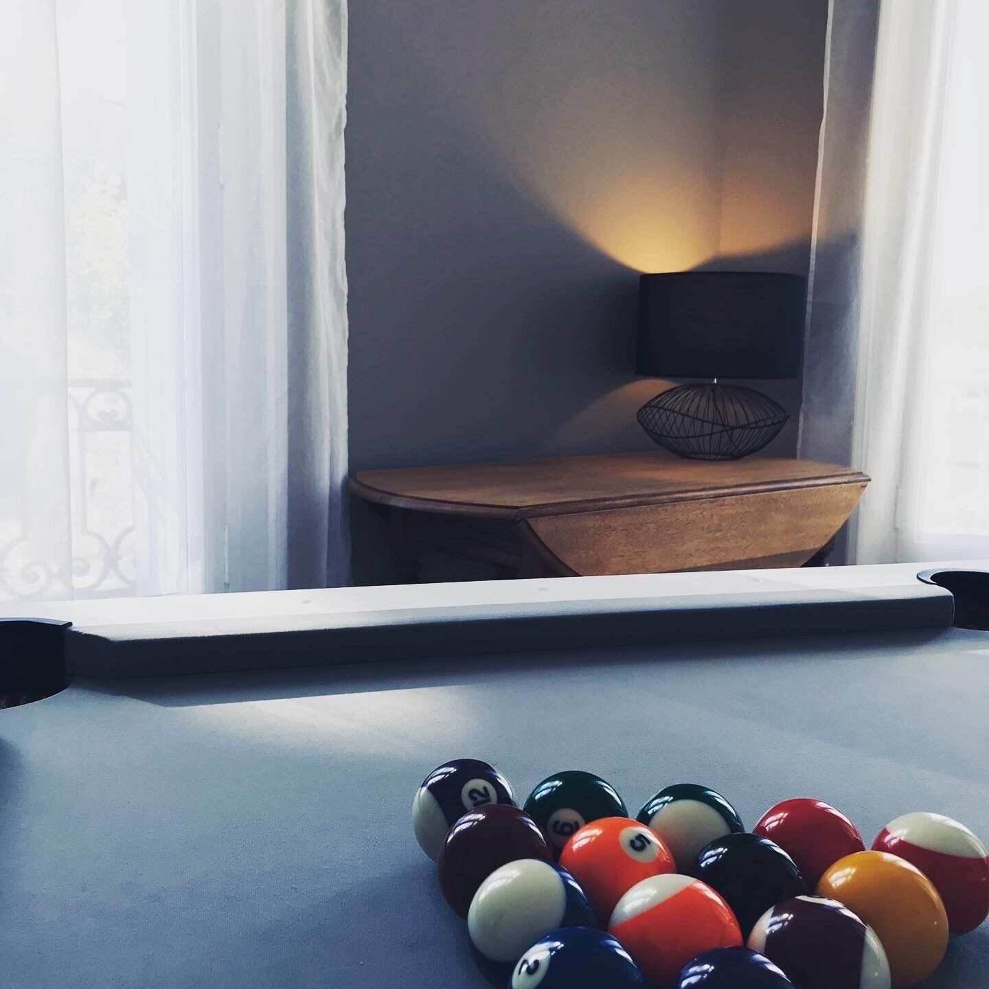 A pool table with billiard balls in a room with a window, a lamp, and a wooden table.