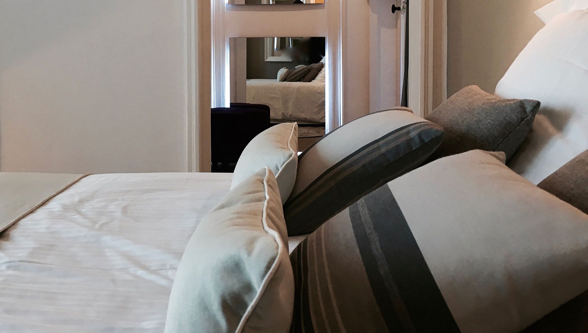 Una cama bien hecha con edredón blanco y almohadas, una mesita de noche con lámpara, y una puerta que da a otra habitación.