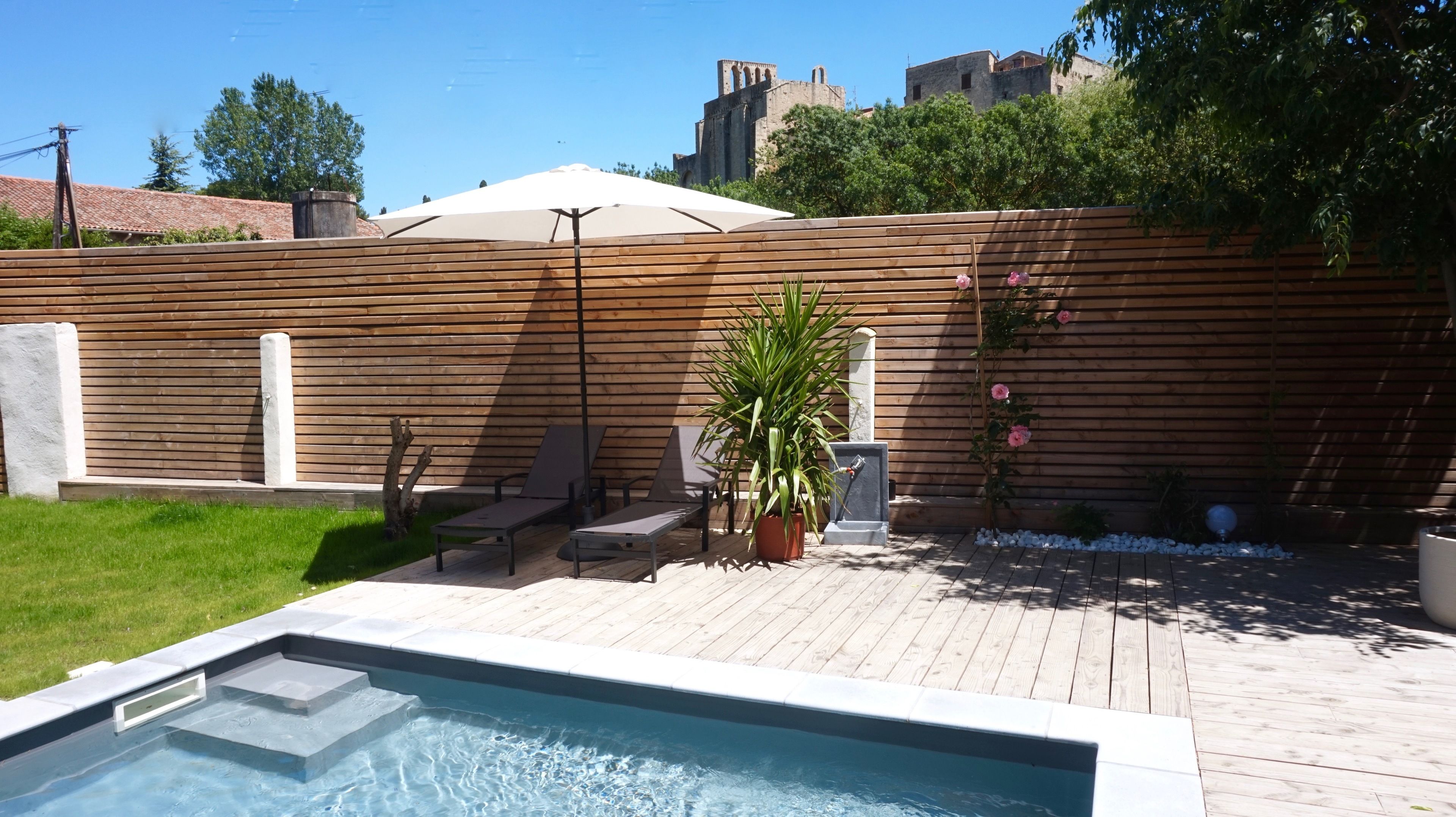A swimming pool with a wooden deck, lounge chairs, a potted plant, and a wooden fence.