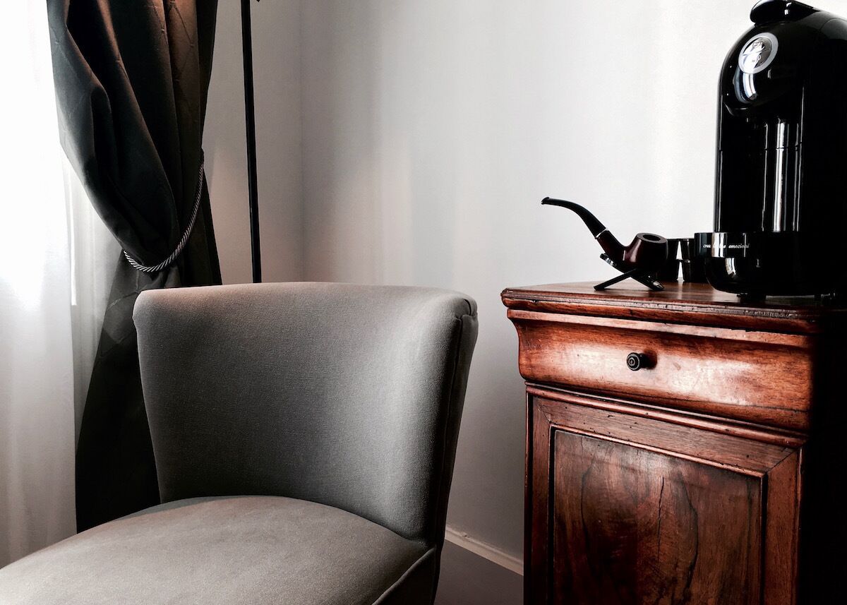 A corner of a room with a grey upholstered chair, a wooden cabinet with a coffee machine on top, and a curtain partially drawn to the side.