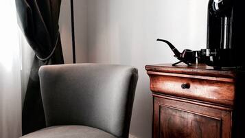 A corner of a room with a grey upholstered chair, a wooden cabinet with a coffee machine on top, and a curtain partially drawn to the side.