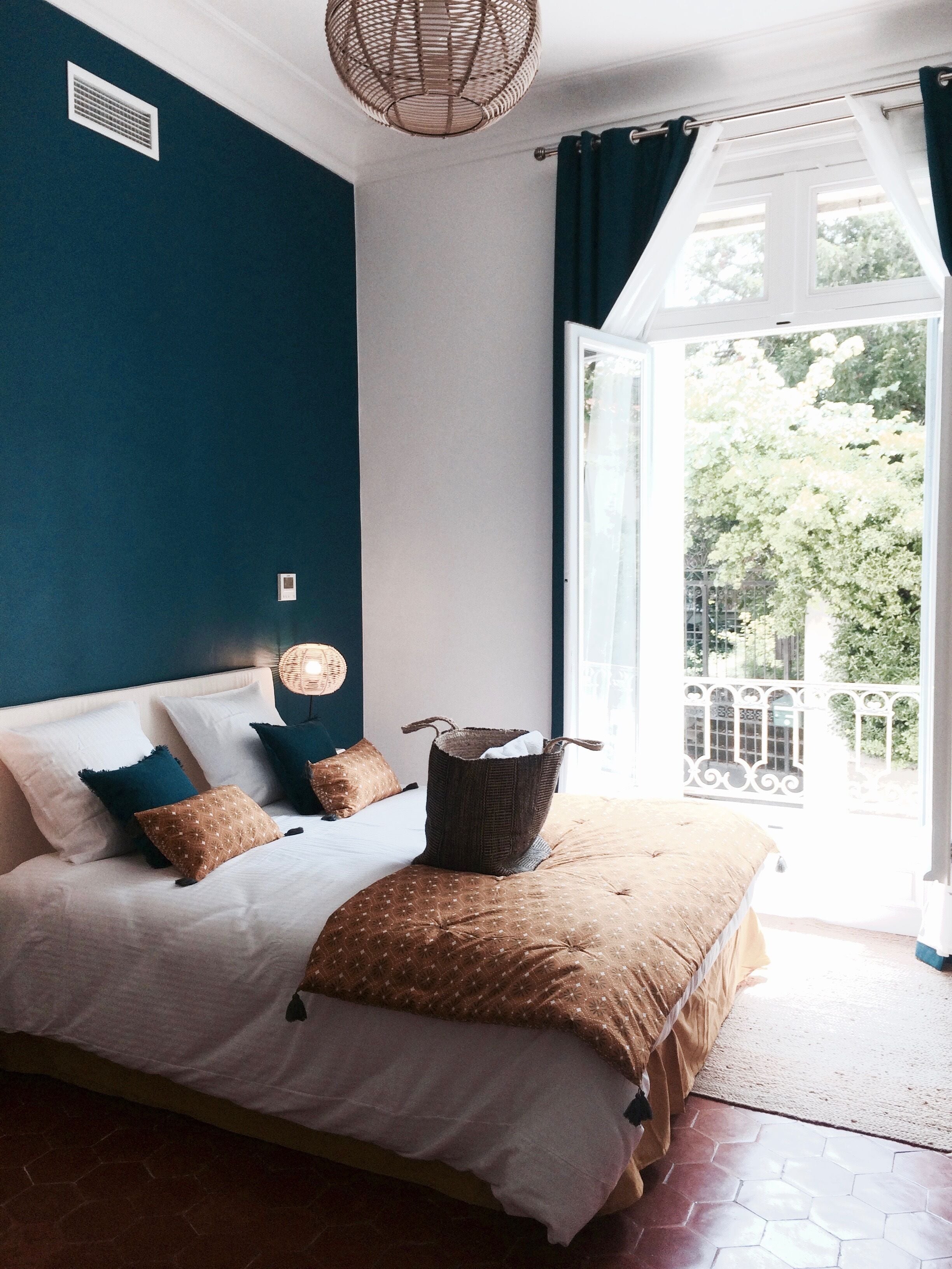 Un dormitorio con cama, una cesta de mimbre, una ventana con cortinas y vista a follaje exterior.
