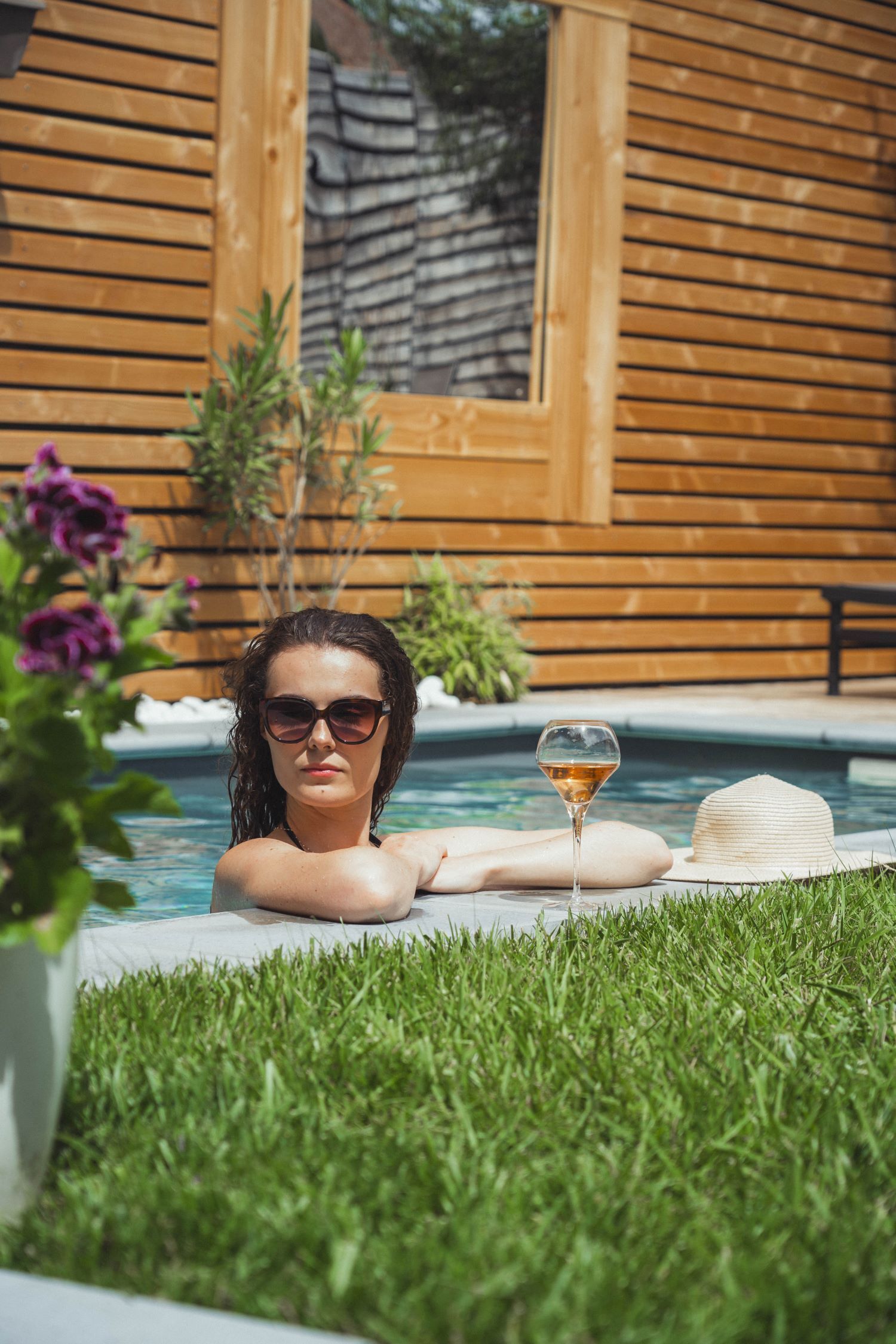 A person relaxing by a pool with a drink and a hat, surrounded by greenery and a wooden building.
