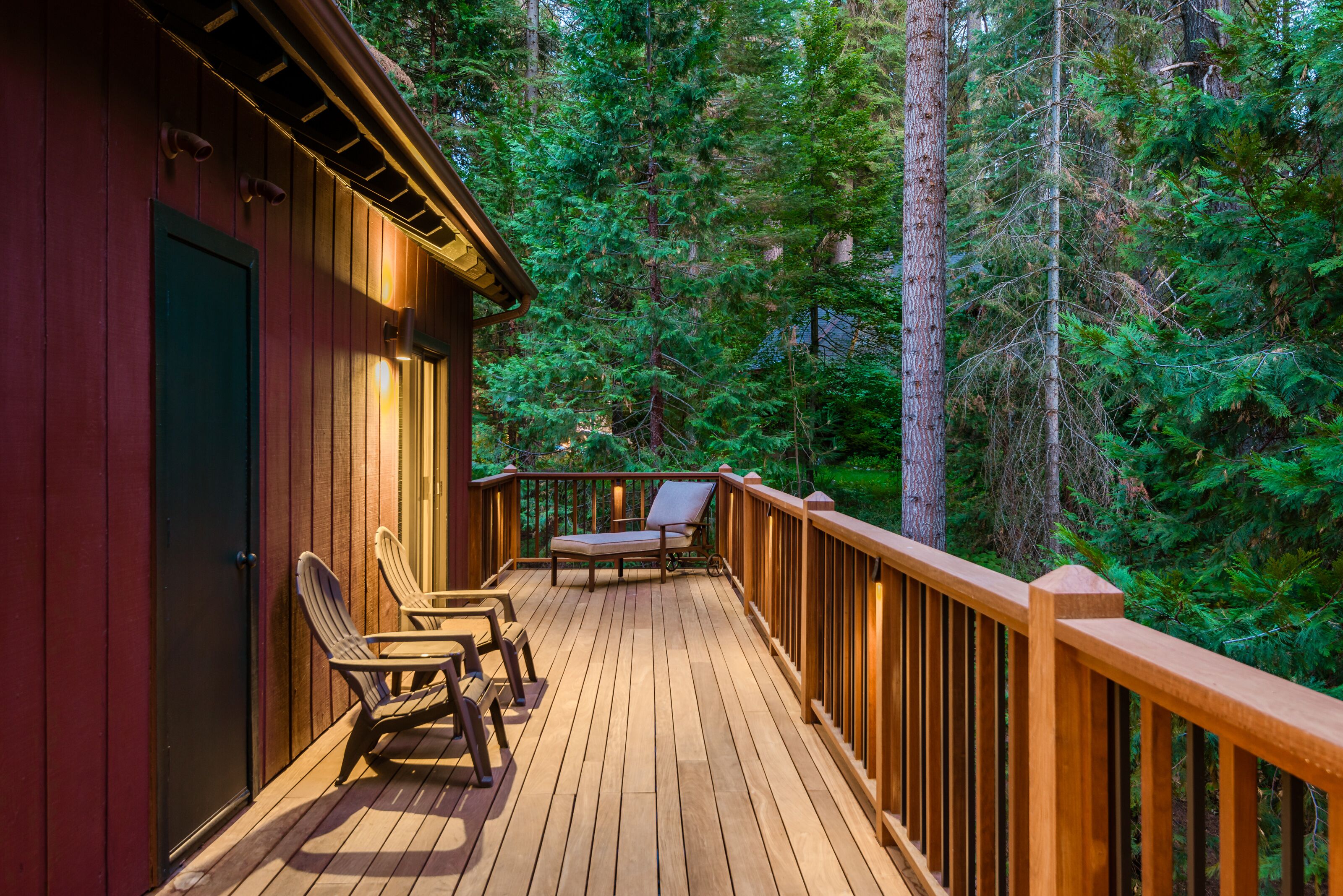 A wooden deck with two lounge chairs and a table, surrounded by a wooden railing, set against a forest backdrop.