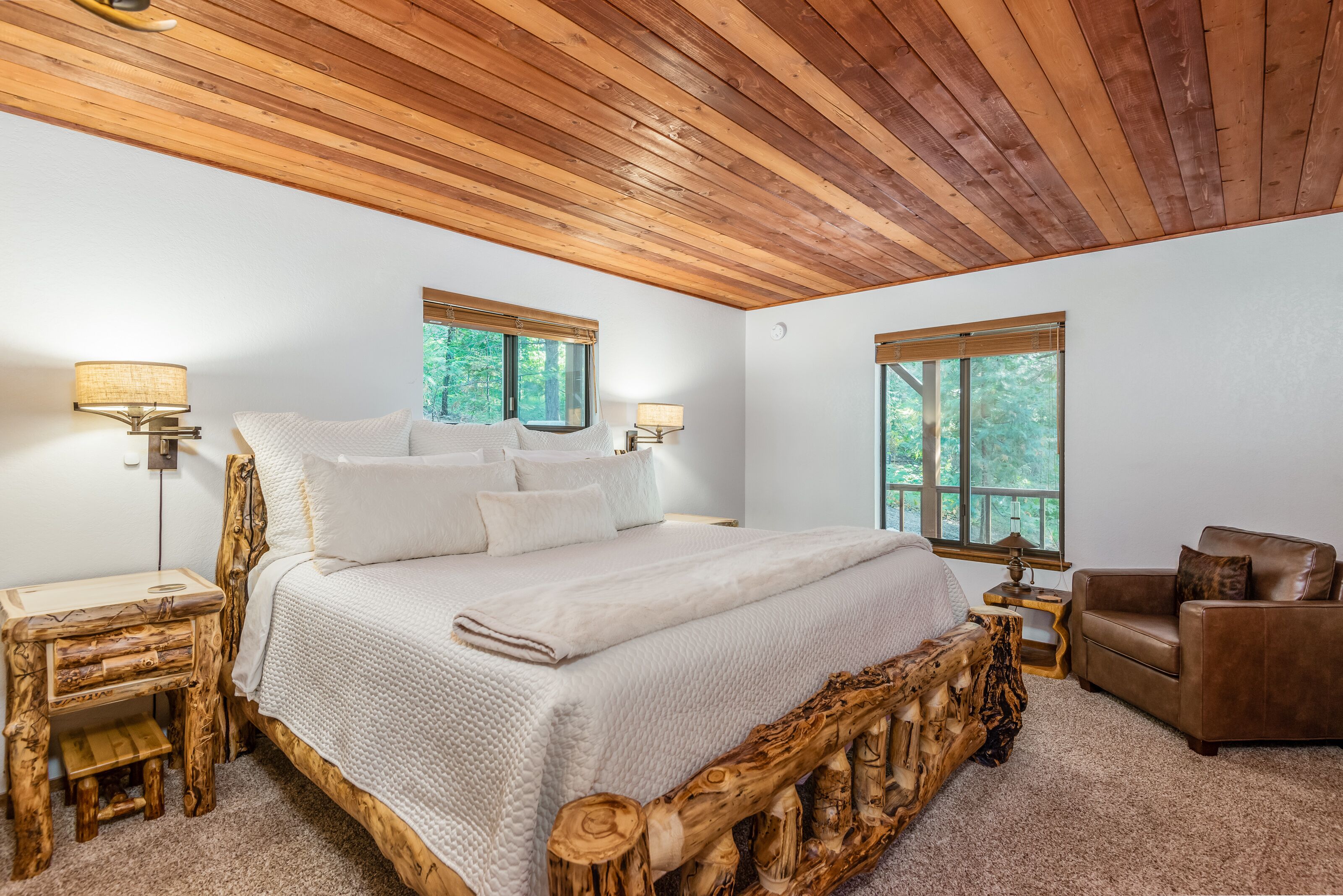 Une chambre à coucher avec une tête de lit en bois, un grand lit avec une literie blanche, deux lampes de chevet, un fauteuil en cuir marron et une fenêtre donnant sur les arbres.