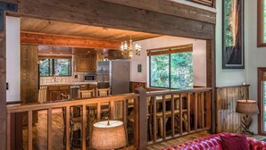 A rustic cabin interior with a kitchen, dining area, and a large window offering a forest view.