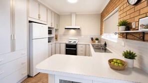 Una cucina moderna con mobili bianchi, forno integrato, piano cottura, lavello e un ripiano con un cesto di mele.