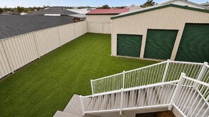 A fenced backyard with artificial grass, a staircase, and two green garage doors. - Seaduced By Minnie - Perfect Getaway (MINNIE WATER)