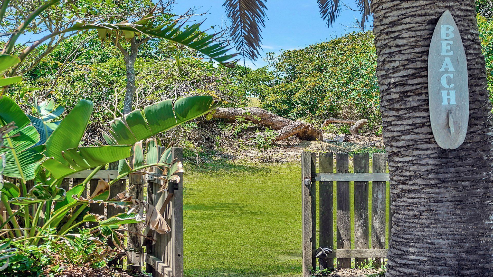A wooden gate with a sign that reads 'BEACH' and a tropical garden with various plants and a palm tree.