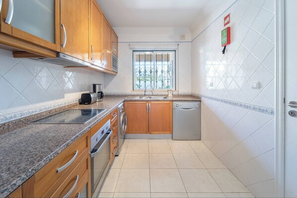 A modern kitchen with wooden cabinets, a stainless steel range hood, a built-in oven, a dishwasher, and a window with a view of greenery.