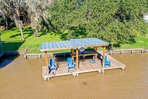 Une terrasse en bois surmontée d’un toit en métal, entourée d’eau, et agrémentée de chaises bleues.