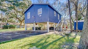 A blue wooden structure with a covered porch, surrounded by trees and a grassy area.