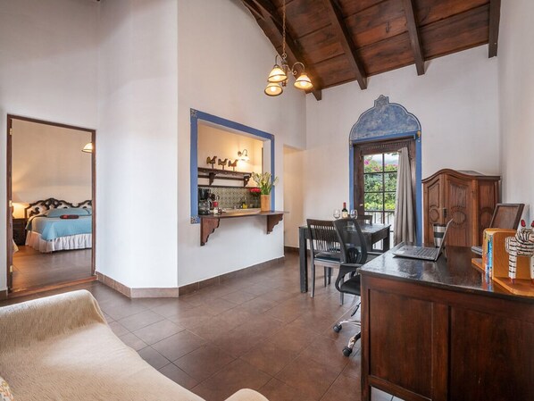 A spacious room with a wooden ceiling, a dining area with a table and chairs, a kitchen area with a counter and shelves, and a view through a window with curtains.