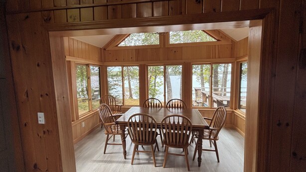Ein Esszimmer mit Holzboden, einem großen Tisch und Stühlen, großen Fenstern mit Blick auf Bäume und Wasser sowie einer Holzbalkendecke.