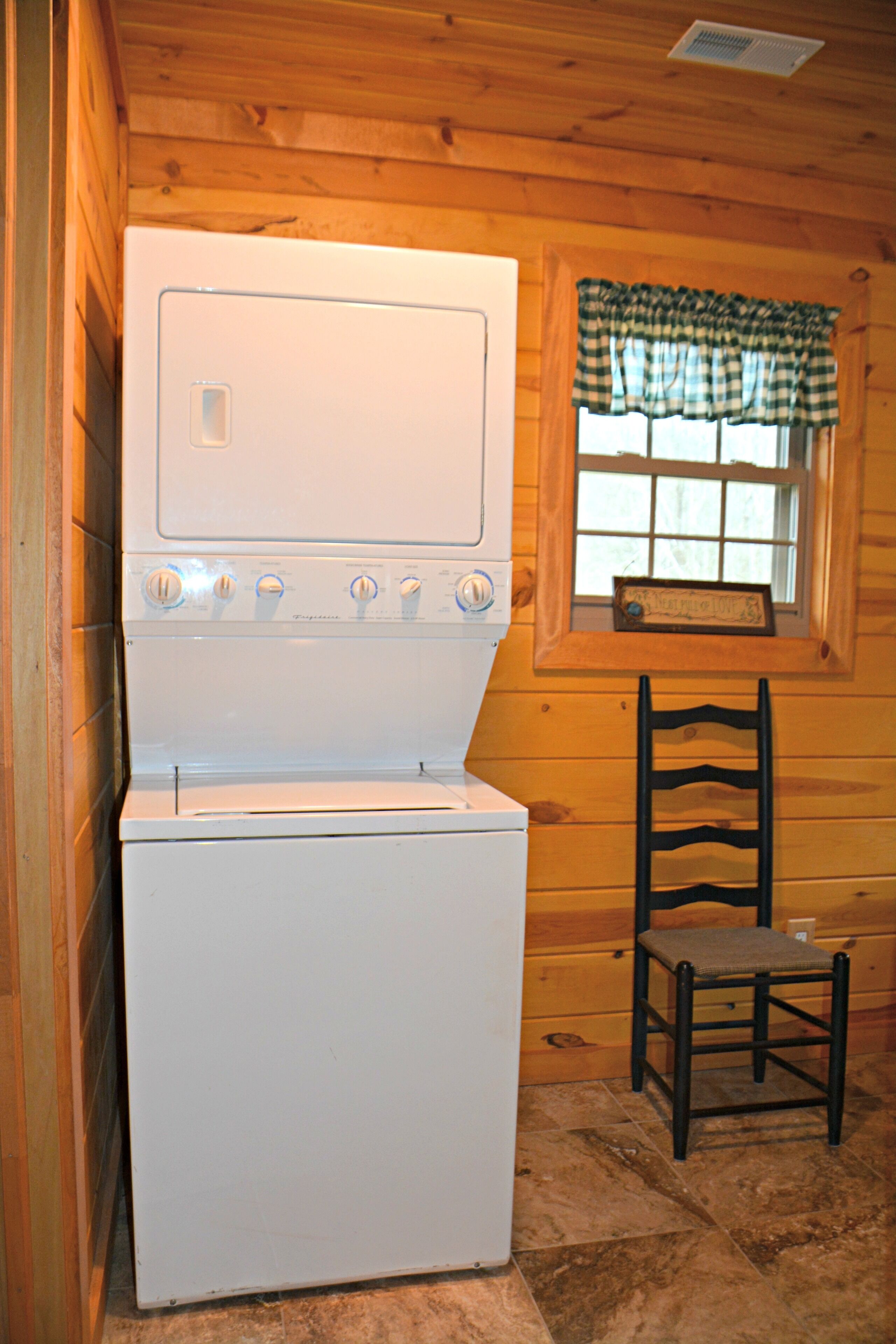 A compact room with a white washer and dryer, a wooden chair, and a window with a checkered valance.