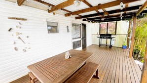 Ein überdachter Außenbereich mit Holztisch und Grill, umgeben von weißen Wänden und einer Holzterrasse.