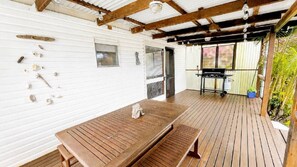 Ein überdachter Außenbereich mit Holztisch und Grill, umgeben von weißen Wänden und einer Holzterrasse.
