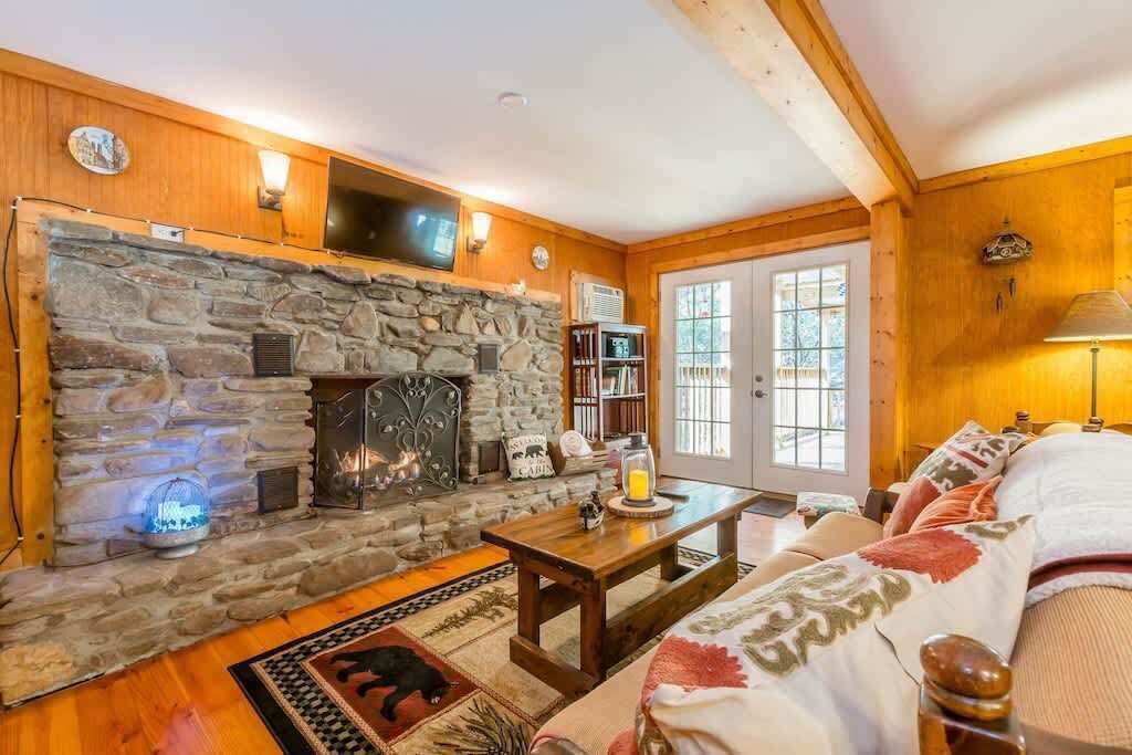 Un salon chaleureux avec une cheminée en pierre, des murs en bois, un téléviseur fixé au-dessus de la cheminée et une grande fenêtre offrant une belle vue.