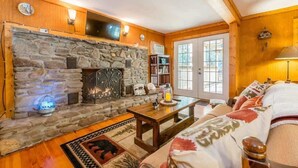 A cozy living room with a stone fireplace, wooden walls, a television mounted above it, and a large window with a view.