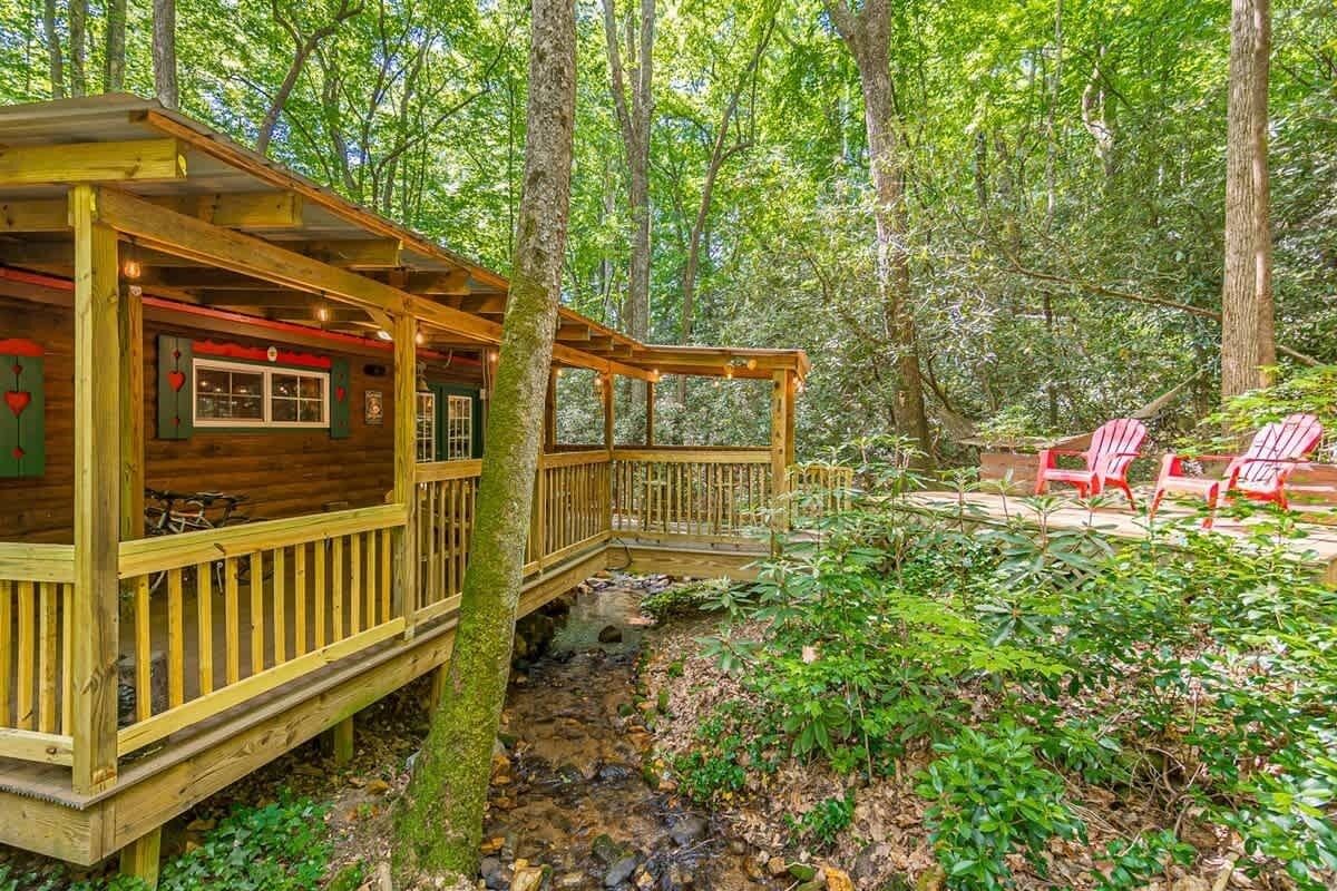 Une cabane dotée d’une terrasse en bois, d’un petit pont et de chaises rouges, le tout situé dans un cadre forestier.