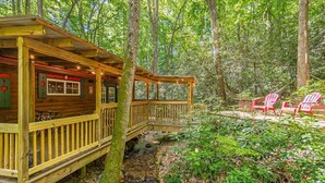 A cabin with a wooden deck, a small bridge, and red chairs in a forest setting.