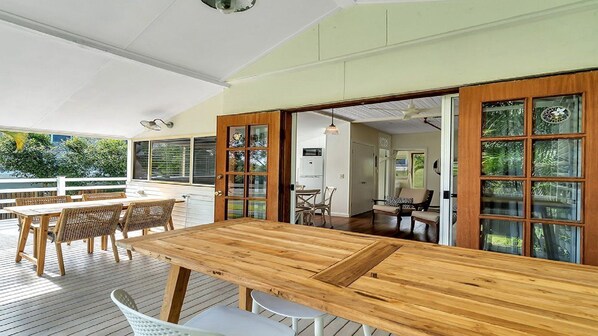 An open-plan living area with a wooden dining table and chairs, a kitchen visible through a glass door, and a view of the outdoors.