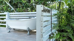 Eine weiße Freiluftbadewanne auf einer Holzterrasse mit Wasserhahn und Duschkopf aus Metall.