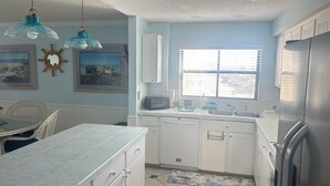 A kitchen with white cabinets, a stainless steel refrigerator, a window with blinds, and a dining area with a table and chairs. - Oceanfront  Sandcastle Condo, South #506 (Ponce Inlet)