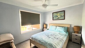 A bedroom with a bed, a ceiling fan, a window with a blind, a bedside table with a lamp, and a painting on the wall.