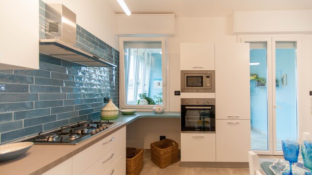 A modern kitchen with a gas stove, microwave, oven, and a window with curtains. There are two baskets on the floor and a table with a tray and glasses. - Entire apartment on the first floor (Piumazzo)