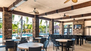 Un espace extérieur aménagé en terrasse, avec des tables et des chaises rondes, une piscine et un coin bar équipé d’un téléviseur.