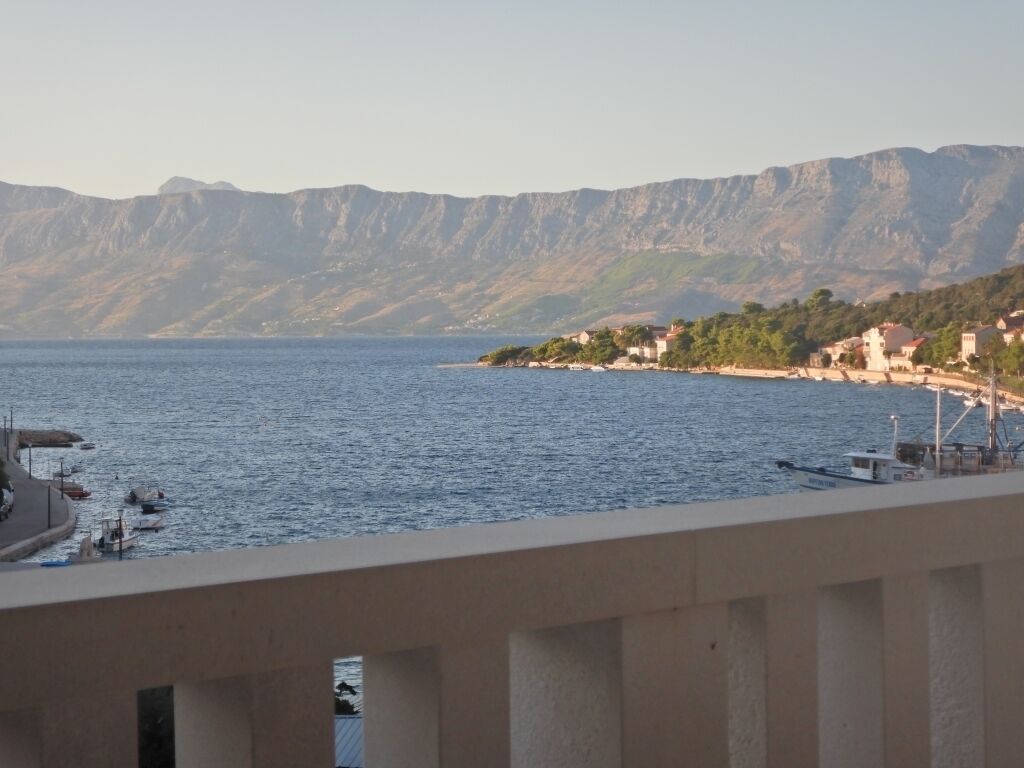 A coastal view with a mountain range in the background, a pier with boats, and buildings along the shore.