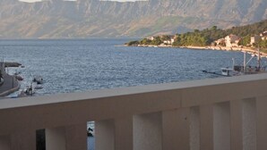 En kustutsikt med ett bergskedja i bakgrunden, en pir med båtar och byggnader längs stranden.