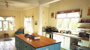 A kitchen with a blue island, a stove, and a sink. There are windows with curtains, a ceiling fan, and a small table with a rug. - Casa Clara | 6BR Villa with Pool & Ocean View | Sleeps 12, Bacolet Tobago (Tobago)