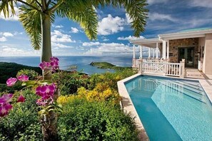 A pool with a view of the ocean, surrounded by lush greenery and a white gazebo.