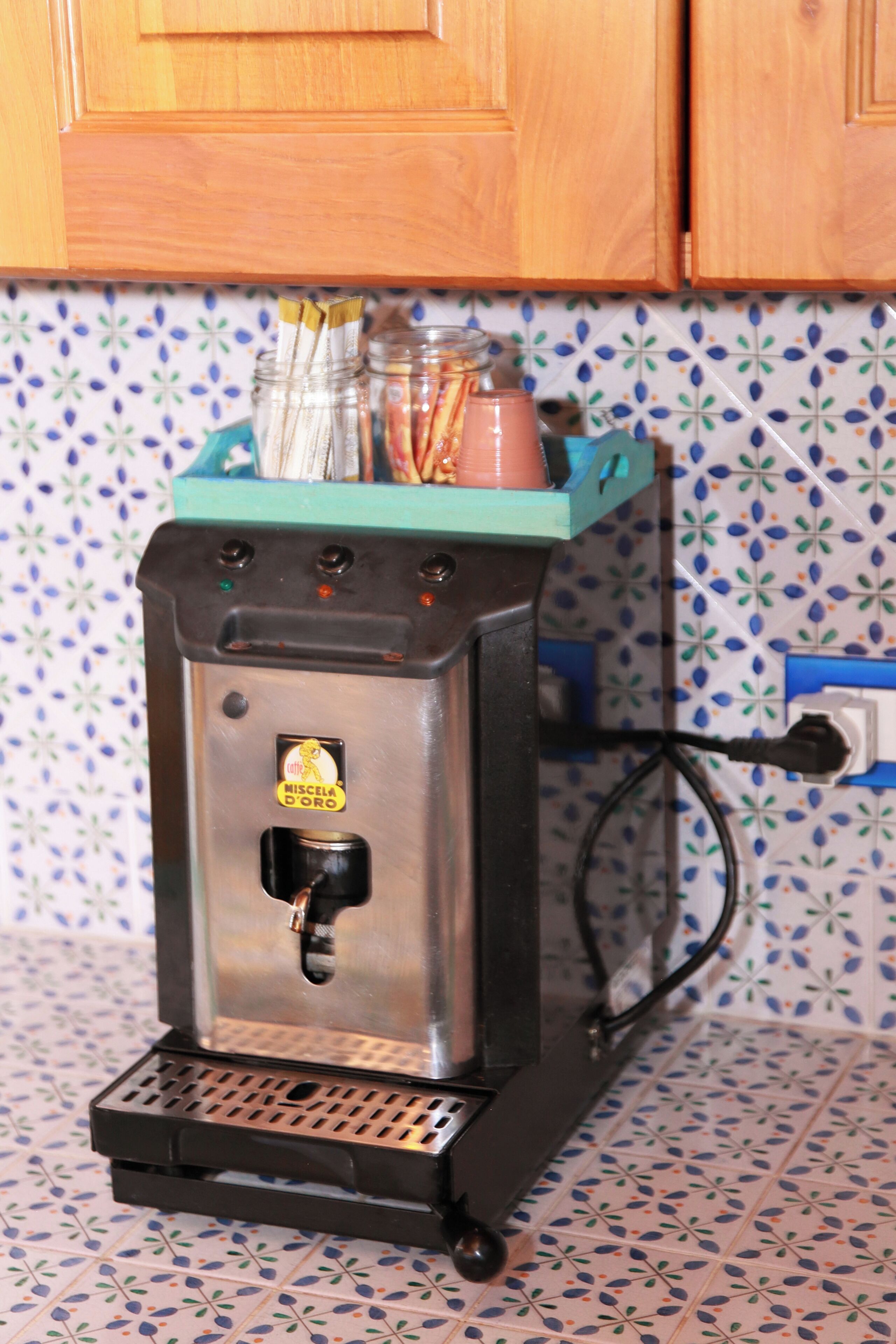 Eine Kaffeemaschine mit Metallgehäuse und schwarzem Sockel, auf einem Fliesenboden mit blau-weißer Musterung.