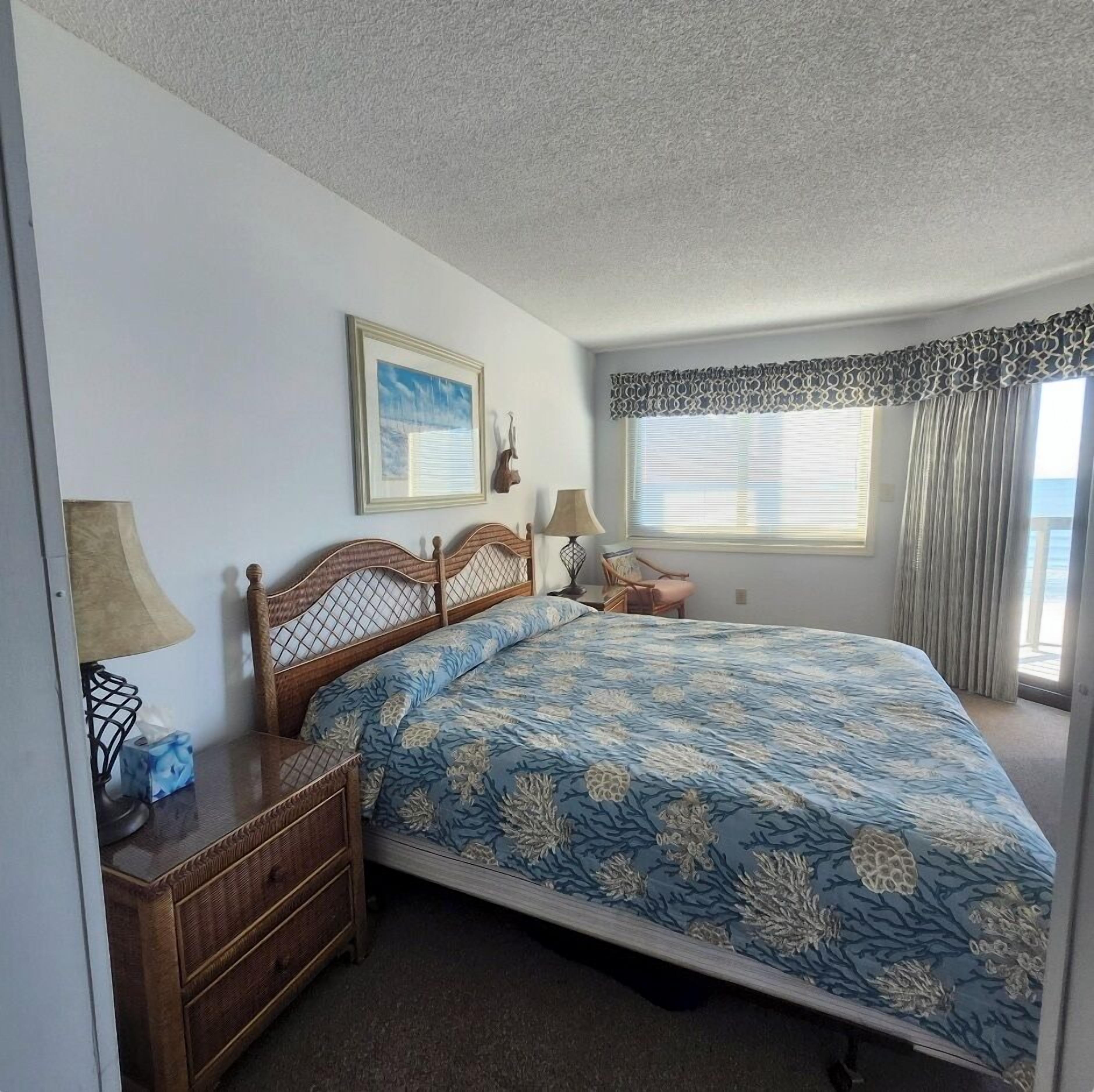 A bedroom with a bed, a nightstand, a lamp, a dresser, a framed picture, and a window with curtains.
