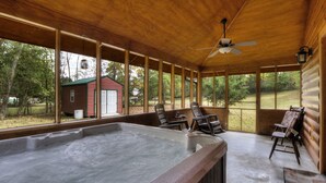 Ein überdachter Außenbereich mit Whirlpool, Holzbalken, Deckenventilator und Blick auf eine rote Holzhütte im Hintergrund.