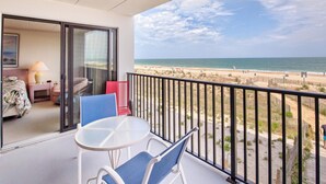 A balcony with a table and chairs, a bed with a floral bedspread, and a view of the beach and ocean.