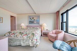 Un dormitorio con cama, dos lámparas de noche, una silla, ventana con vista a la playa y un cuadro en la pared.