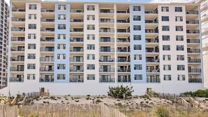 A multi-story residential building with balconies, a sandy ground level, and a clear sky.