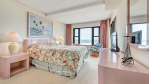 A hotel room with a bed, two bedside tables, a chair, a large window with a view of the sea, and a painting on the wall.