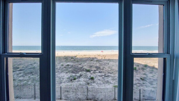 Une vue à travers trois fenêtres sur une plage, avec une clôture en bois au premier plan et une plage de sable où l’on aperçoit quelques personnes au loin.